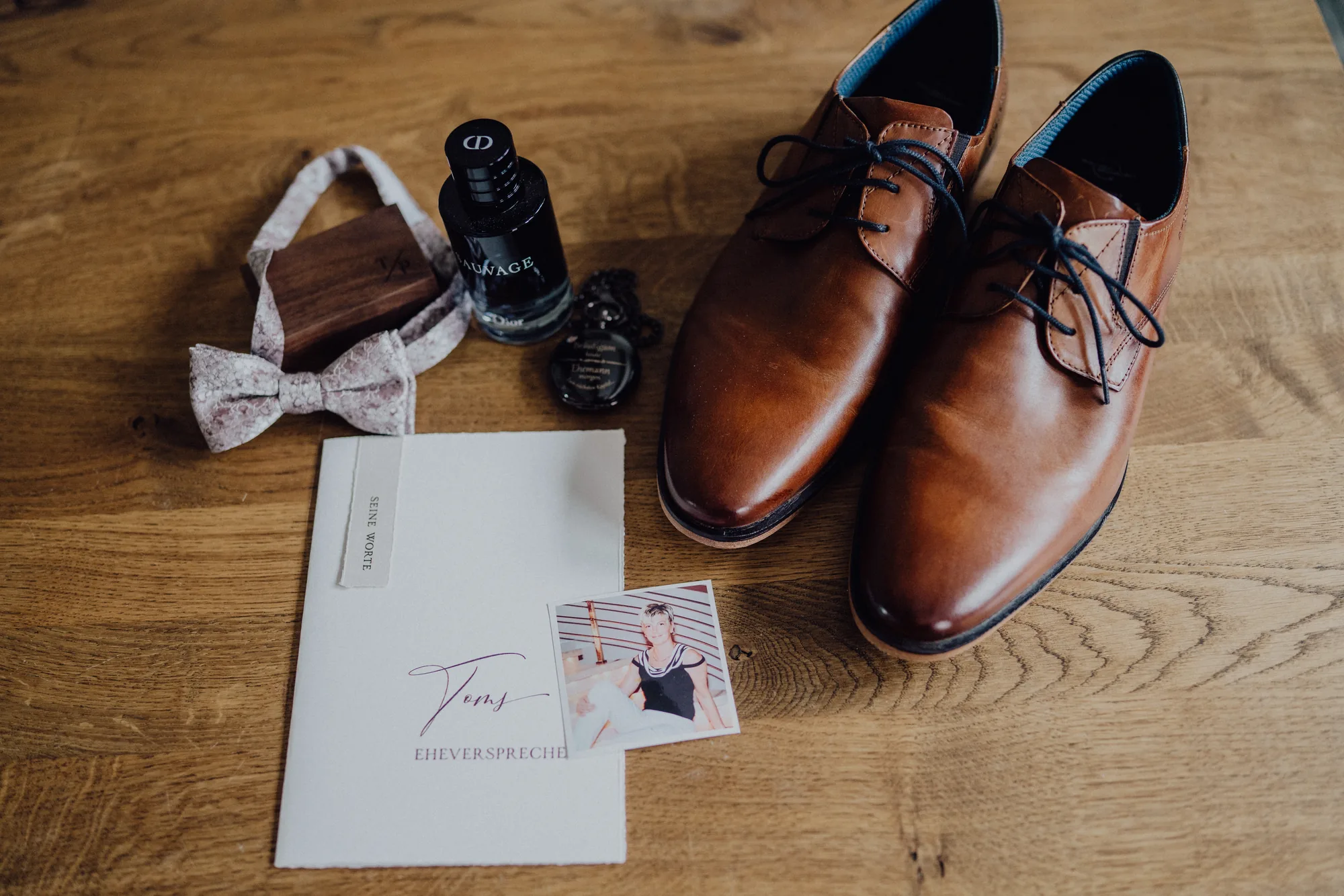Toms Fliege, Parfum, Lederschuhe und sein Eheversprechen auf einem Holztisch – Hochzeit Bella & Tom, Gut Immenhof, Malente