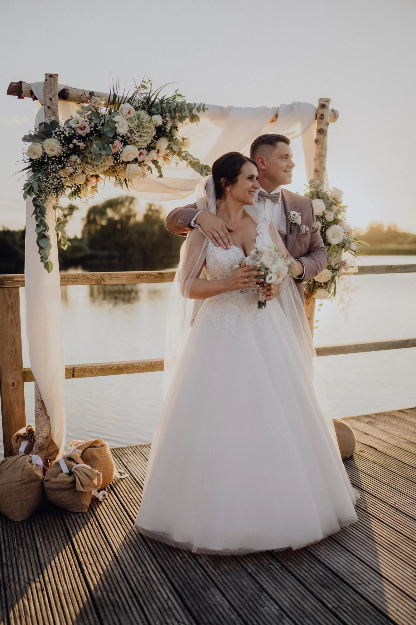 Bella und Tom am blumengeschmückten Traubogen auf dem Steg am See – Hochzeit Bella & Tom, Gut Immenhof, Malente