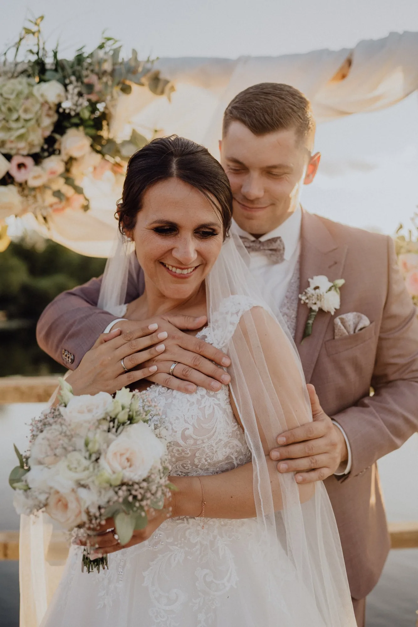 Tom umarmt Bella von hinten, ihre Hände mit den Eheringen liegen übereinander – Hochzeit Bella & Tom, Gut Immenhof, Malente