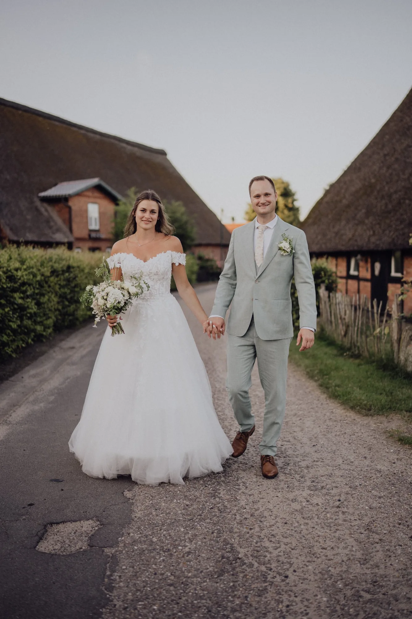 Franzi und Tim gehen Hand in Hand über den Dorfweg zwischen den Reetdachhäusern – Hochzeit Franzi & Tim, De Ole Schüün, Hüttblek