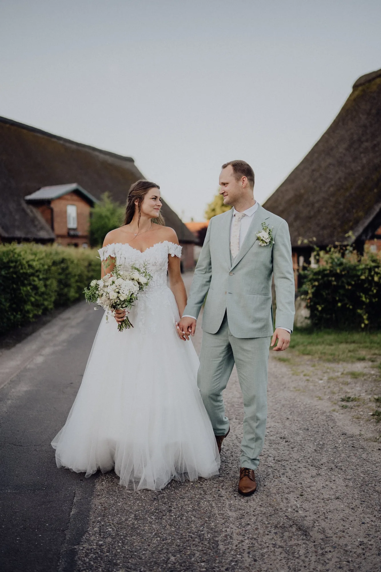 Franzi und Tim schauen sich beim Gehen an, Reetdächer im Hintergrund – Hochzeit Franzi & Tim, De Ole Schüün, Hüttblek